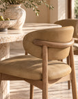 A close-up of a modern dining chair with a curved backrest upholstered in warm tan leather or suede, supported by a light natural wood frame, set in a sunlit dining room next to a stone table.