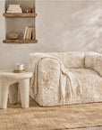 A cream-colored, shaggy faux-fur armchair styled in a warm, rustic room with a matching woven rug, a small stone side table, and a vase of dried pampas grass.
