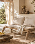 Karoo Sofa two-seater with a natural timber frame and light upholstered cushions, styled in a covered outdoor lounge with soft linen curtains and garden views.