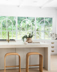 Uniqwa Collections Elula Counter Stools in natural oak featured in a contemporary white kitchen with curved travertine benchtop and rustic accents