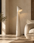 A modern sculptural floor lamp with a textured off-white finish stands in a sunlit minimalist living room next to a cream sofa and a large stone planter.
