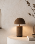 Small Samrah table lamp with textured brown paper mache and dome shade casting a warm ambient glow in a natural styled interior.