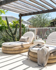 Two Satori Outdoor Sun Loungers with natural woven bases and cream cushions beneath a rustic timber pergola, surrounded by lush coastal garden greenery.