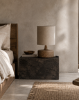 Zaya table lamp with a sculptural textured base and natural fabric shade styled on a dark stone bedside table in a warm and tranquil bedroom.