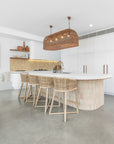 Uniqwa Collections Tula Bar Chairs featured in a modern coastal kitchen with white cabinetry, curved island bench, woven pendant lights, and polished concrete floors