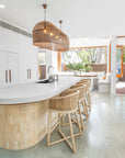 Uniqwa Collections Tula Bar Chairs featured in a modern coastal kitchen with white cabinetry, curved island bench, woven pendant lights, and polished concrete floors.