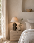 Liwa Table Lamp on a timber bedside table, featuring a textured sculptural base and tapered shade in a softly lit bedroom setting