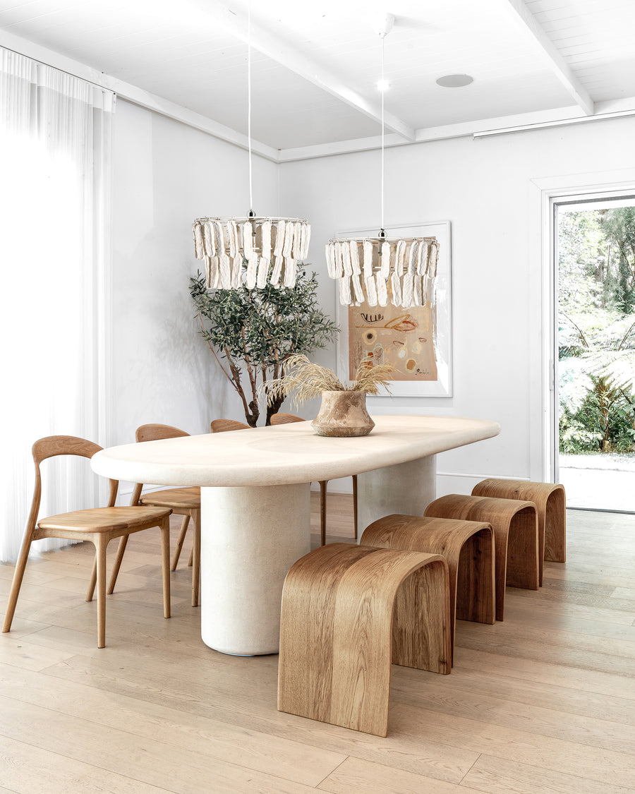 A contemporary dining space featuring a sculptural oval dining table, curved wooden stools, and minimalist wooden chairs, complemented by soft pendant lighting and organic décor