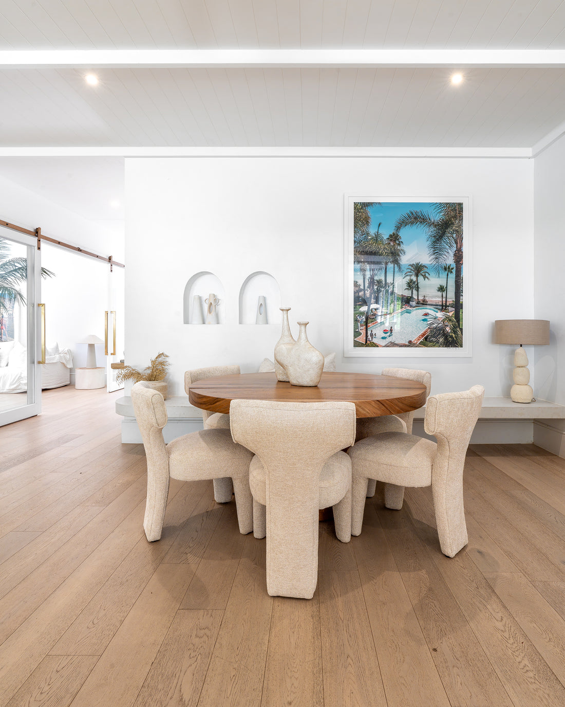 A sophisticated dining setup featuring plush boucle dining chairs surrounding a round wooden table, complemented by minimalist décor and a coastal-inspired art piece in a bright, airy space