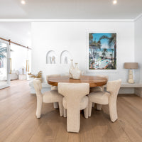 A sophisticated dining setup featuring plush boucle dining chairs surrounding a round wooden table, complemented by minimalist décor and a coastal-inspired art piece in a bright, airy space