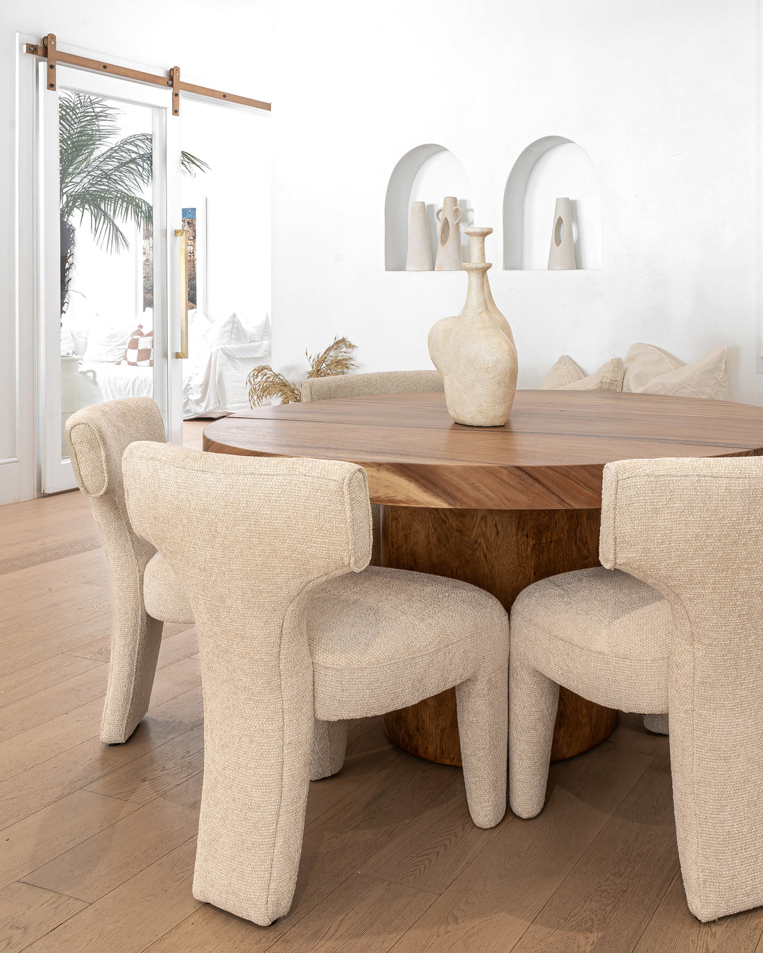 A close-up view of elegant boucle dining chairs around a round wooden table, featuring sculptural vases and neutral-toned décor in a bright, modern space.