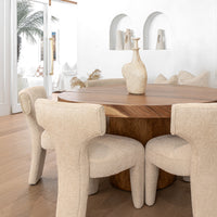 A close-up view of elegant boucle dining chairs around a round wooden table, featuring sculptural vases and neutral-toned décor in a bright, modern space.