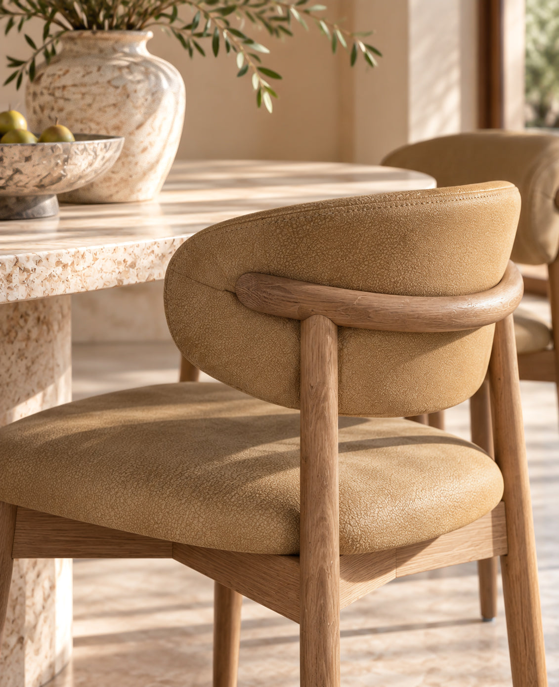 A close-up of a modern dining chair with a curved backrest upholstered in warm tan leather or suede, supported by a light natural wood frame, set in a sunlit dining room next to a stone table.