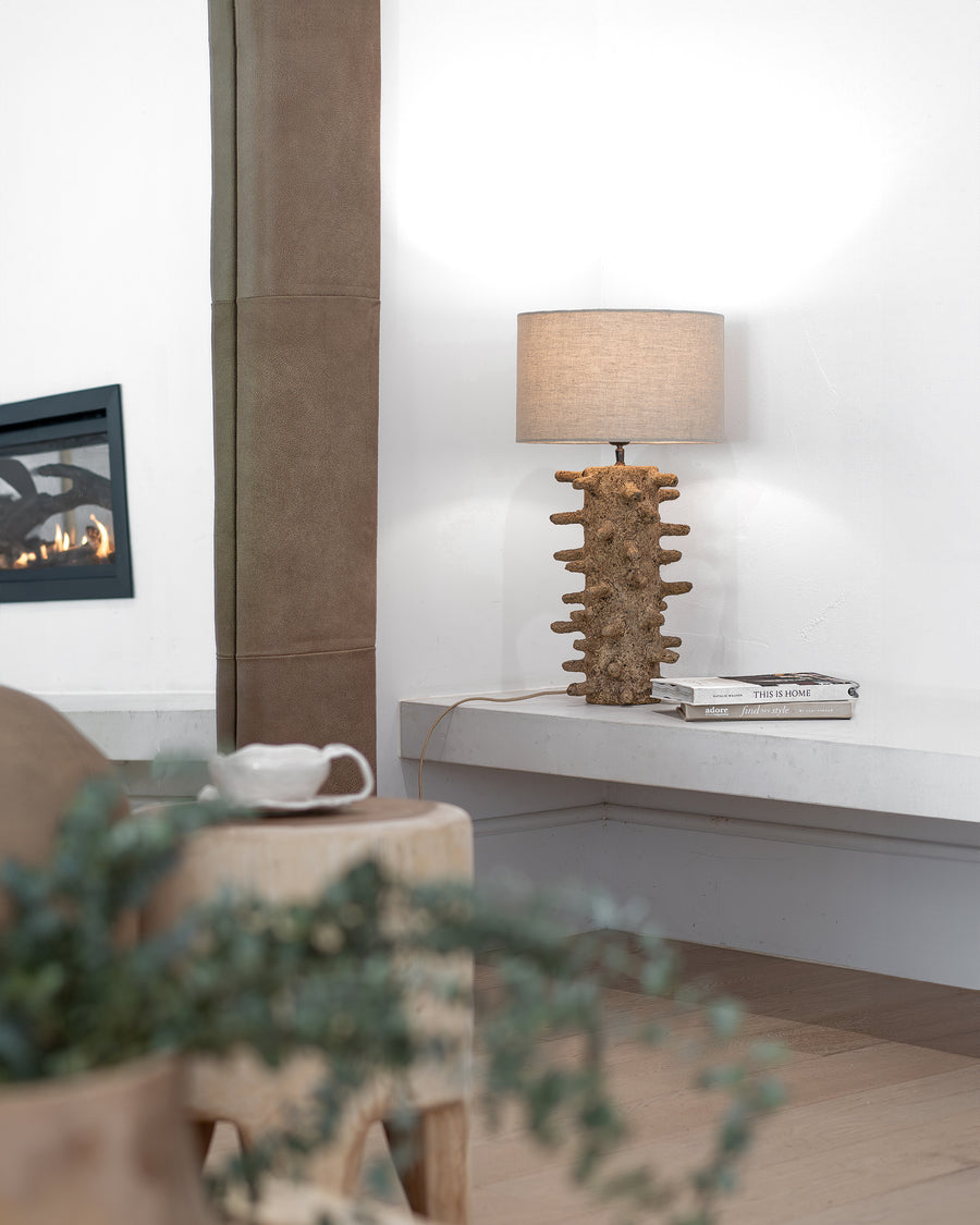 Coral-textured table lamp with a neutral fabric shade styled on a white floating console, complemented by a raw side table and lounge, creating a warm and organic living space with a fireplace in the background