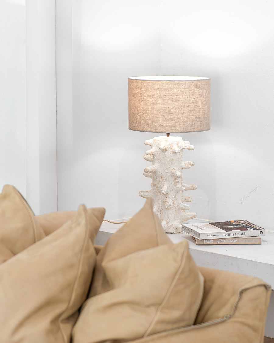 Uniqwa Collections Coral-inspired textured table lamp with a neutral fabric shade styled on a white console beside a tan leather lounge with layered cushions, creating a warm and organic living space