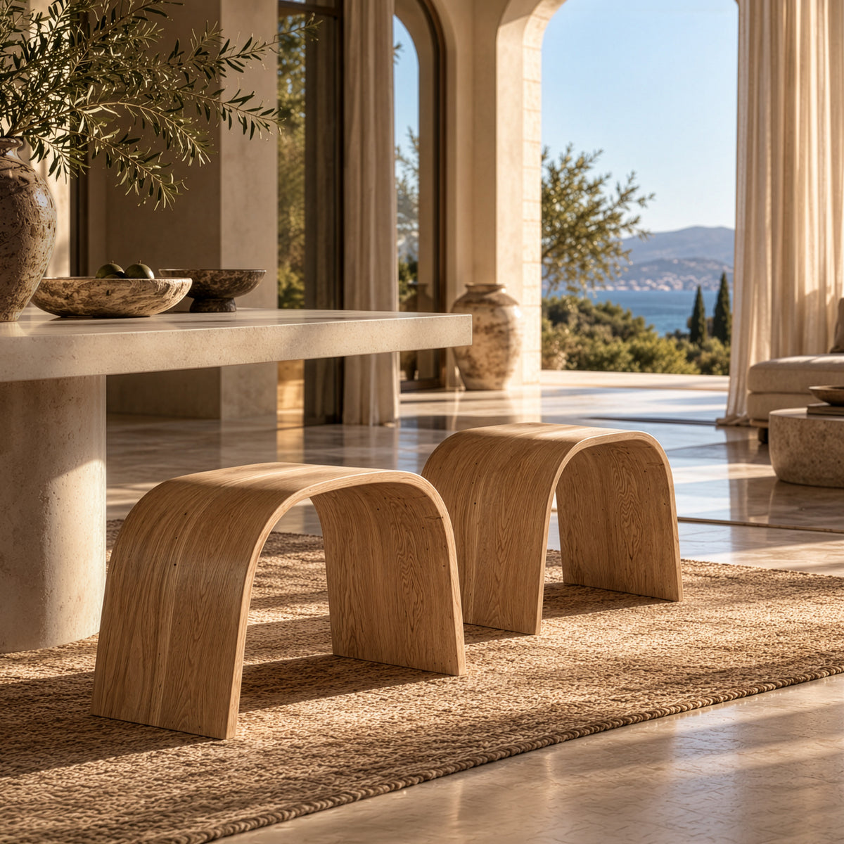 Elula Stool Wide in natural timber, featuring a curved arch form and visible wood grain, styled beneath a stone dining table in a coastal interior.
