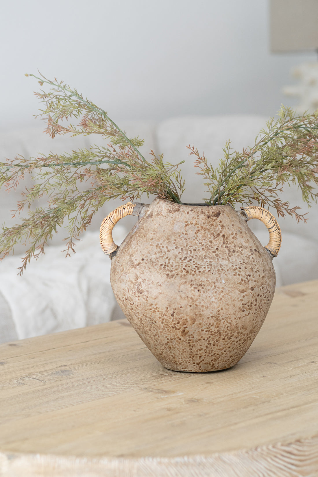 A handcrafted ceramic vase with a weathered texture and rattan-wrapped handles, styled with delicate greenery on a natural wood coffee table. A perfect blend of organic materials and earthy tones for a relaxed, refined aesthetic