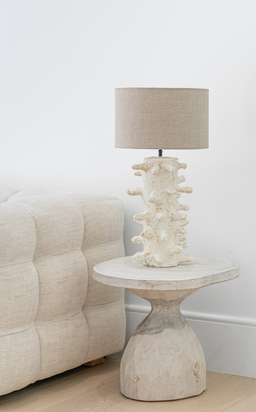 A beautifully handcrafted wooden side table paired with an organic sculptural lamp, featuring a textured natural finish. Styled next to a plush linen sofa, this setup exudes earthy sophistication and timeless elegance