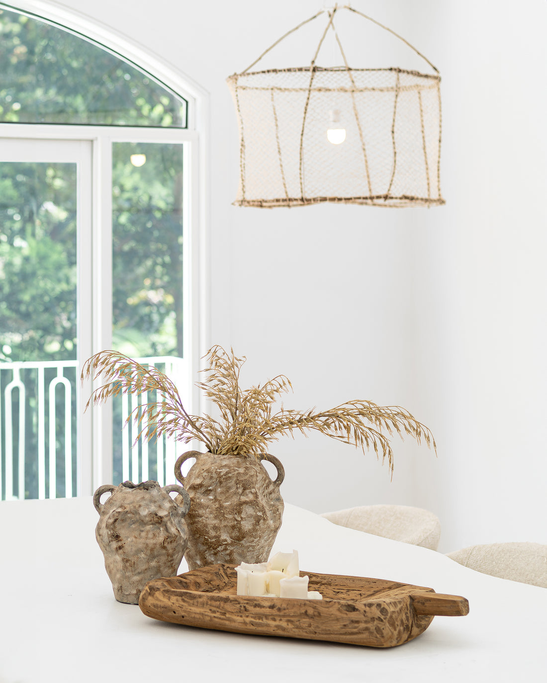 Uniqwa Colletions handwoven net pendant light hanging above a white dining table styled with Ukoko Vase, and a rustic carved naga wooden tray with candles, creating a natural and textural interior aesthetic.