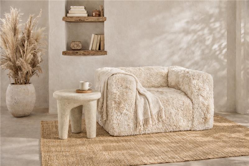 A cream-colored, shaggy faux-fur armchair styled in a warm, rustic room with a matching woven rug, a small stone side table, and a vase of dried pampas grass.