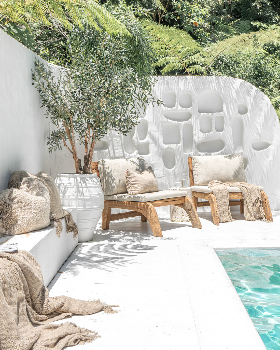 Sunlit poolside oasis featuring rustic timber lounge chairs with soft neutral cushions, a sculptural white wall, and lush greenery, creating a tranquil and inviting outdoor retreat