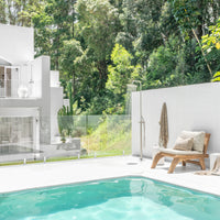 Serene outdoor poolside retreat featuring a natural timber lounge chair with neutral cushions, complemented by lush greenery and a minimalist white backdrop, creating a tranquil and stylish alfresco setting