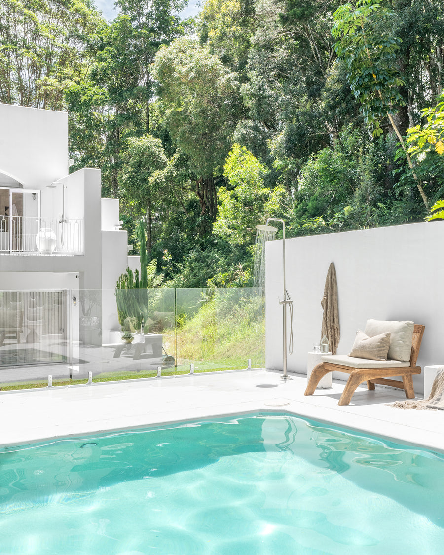 Serene outdoor poolside retreat featuring a natural timber lounge chair with neutral cushions, complemented by lush greenery and a minimalist white backdrop, creating a tranquil and stylish alfresco setting