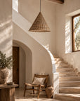 A rustic woven wicker pendant light hangs in a sunlit interior with plaster walls, a stone staircase, a wooden chair, and a large vase on a side table.