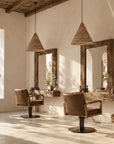 A rustic salon interior with two brown leather barber chairs, large wood-framed mirrors, and woven pendant lights in a warm, sunlit room with plaster walls and exposed wooden ceiling beams.