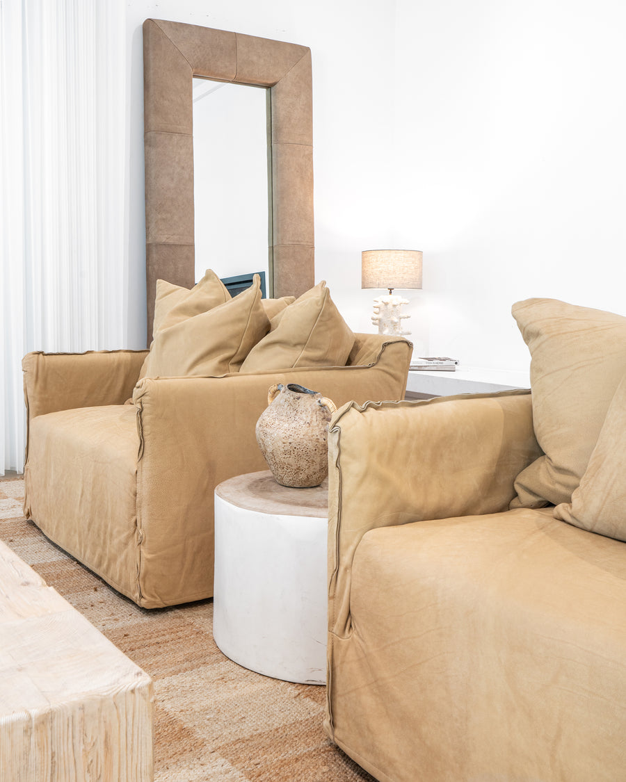 Leather sofa with oversized cushions styled with a vase on a round trunk side table, complemented by a textured rug and a coral-inspired table lamp in a refined organic living space