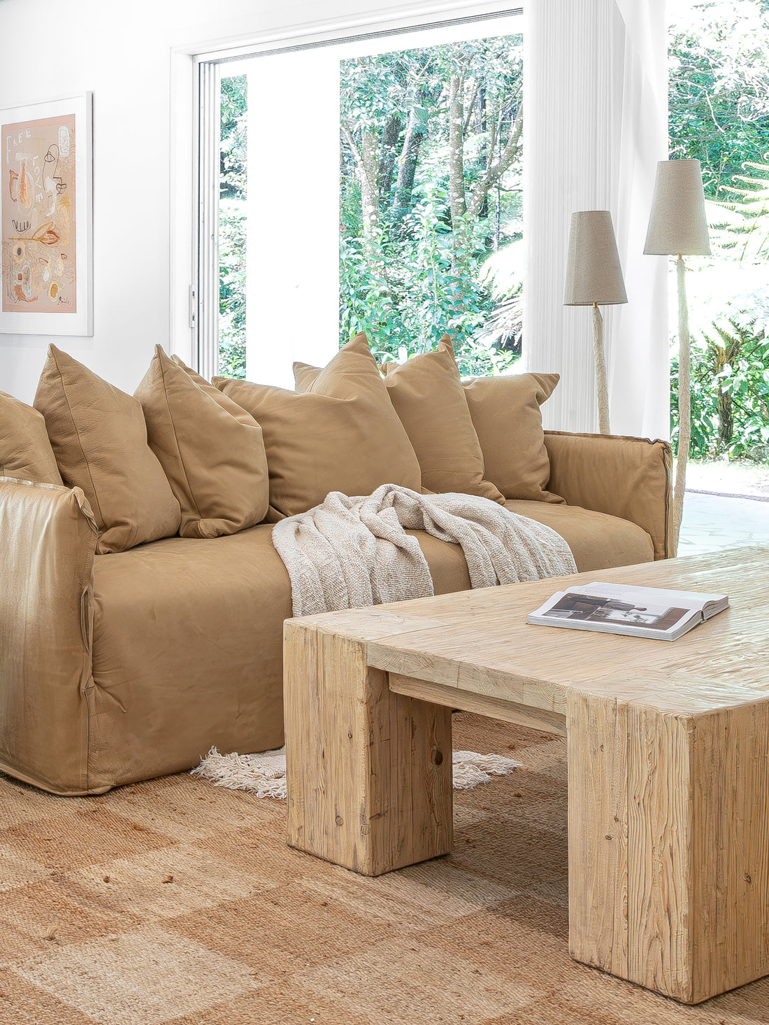 Leather sofa styled with oversized cushions and a textured throw, paired with a reclaimed wood coffee table on a woven jute rug, complemented by sculptural floor lamps in a serene organic living room