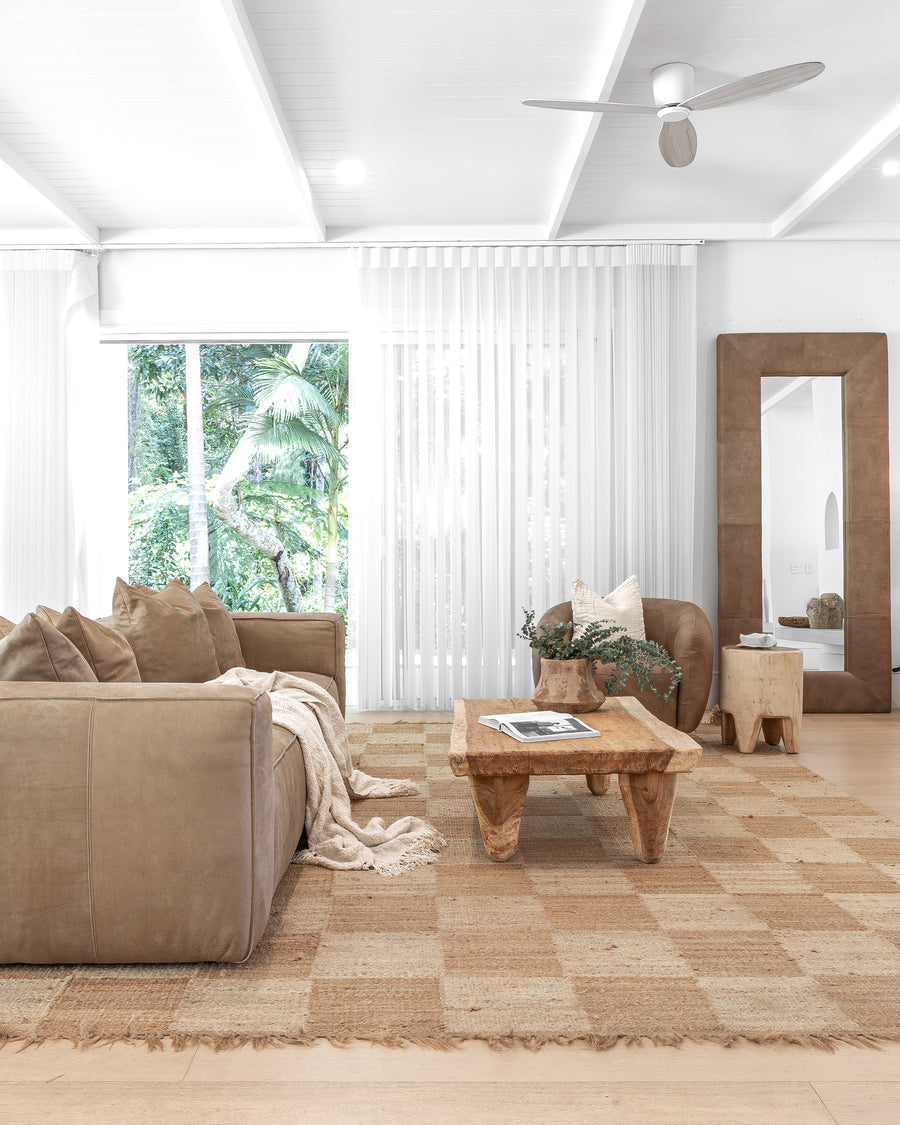 Modern living room featuring a leather sofa, handcrafted timber coffee table, and woven jute rug, complemented by a leather-framed mirror and minimalist decor in a serene, natural setting