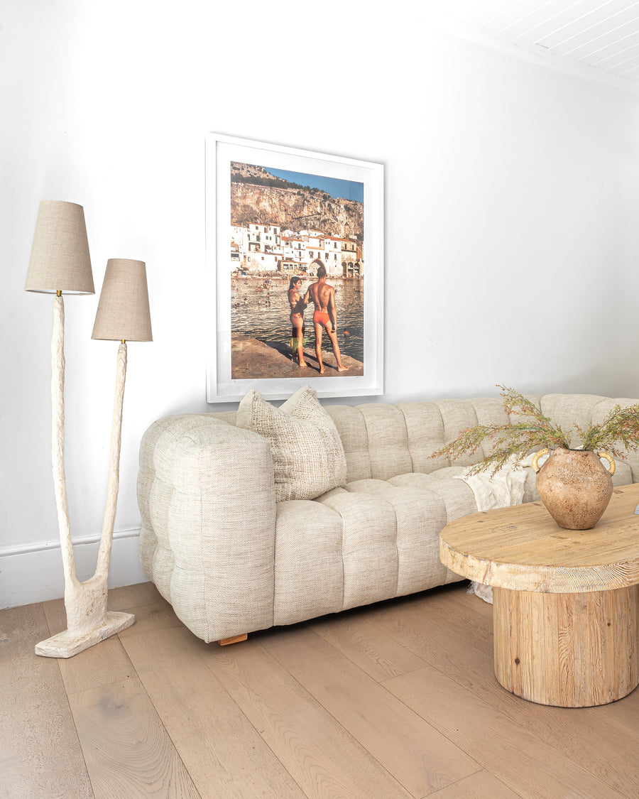 Fabric tufted sofa styled with a reclaimed wood coffee table and sculptural floor lamps, complemented by organic textures and a coastal-inspired framed artwork in a serene modern living space