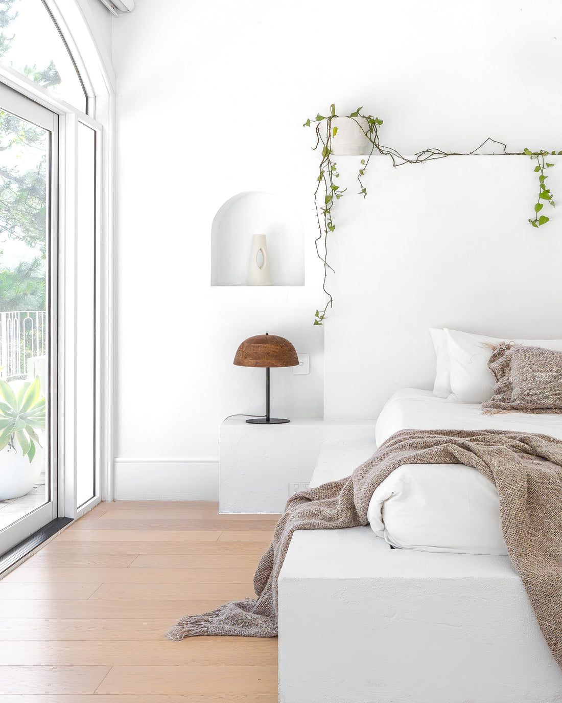 Minimalist bedroom featuring a white plaster built-in bed with soft earthy-toned bedding, complemented by the Sahra Table Lamp and cascading greenery for a serene and organic aesthetic