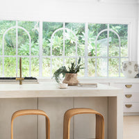 Minimalist kitchen island with a smooth stone finish, styled with curved wooden bar stools, brass tapware, and a rustic ceramic vase, set against large arched windows overlooking lush greenery