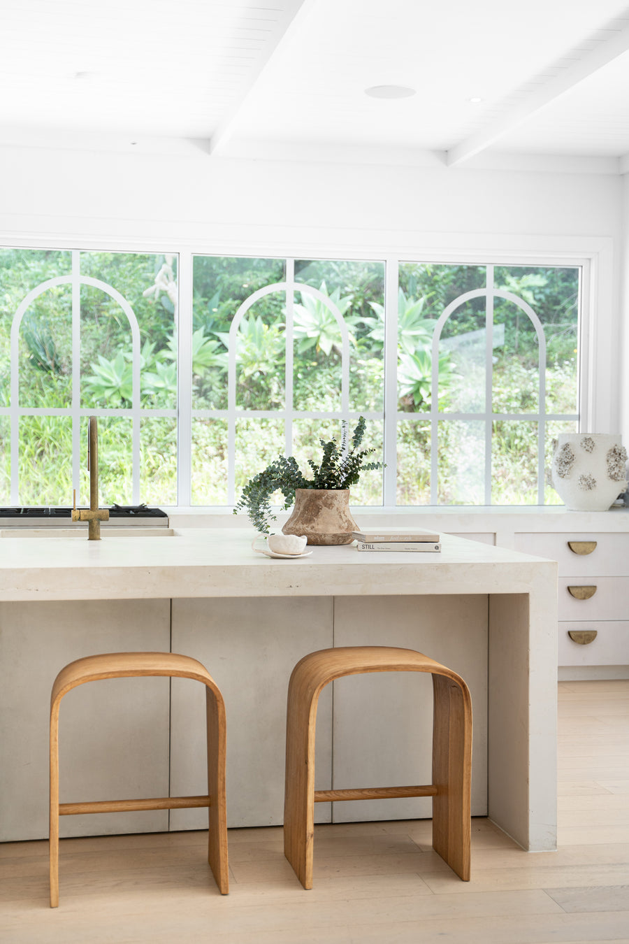 Minimalist kitchen island with a smooth stone finish, styled with curved wooden bar stools, brass tapware, and a rustic ceramic vase, set against large arched windows overlooking lush greenery