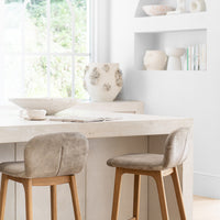 Elegant kitchen setting featuring leather-upholstered wooden bar stools, a natural stone countertop, and minimalist built-in shelving adorned with artisanal ceramics and decor, creating a refined and organic aesthetic