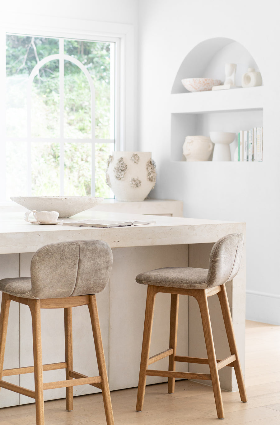 Elegant kitchen setting featuring leather-upholstered wooden bar stools, a natural stone countertop, and minimalist built-in shelving adorned with artisanal ceramics and decor, creating a refined and organic aesthetic