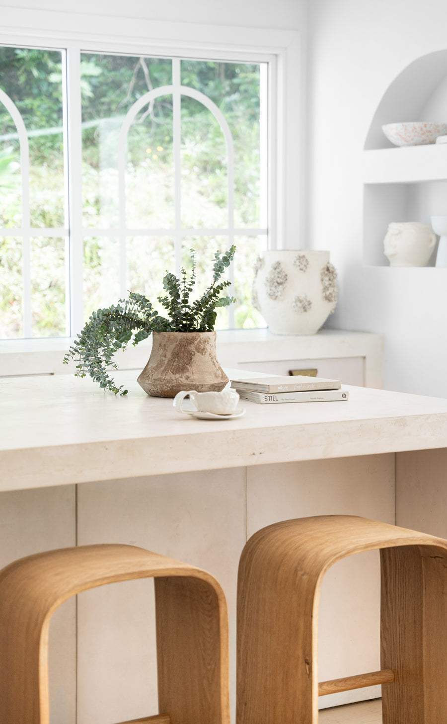 Minimalist kitchen space featuring sculptural wooden bar stools, a natural stone countertop, and earthy decor with organic ceramic vases and fresh greenery, creating a serene and inviting atmosphere