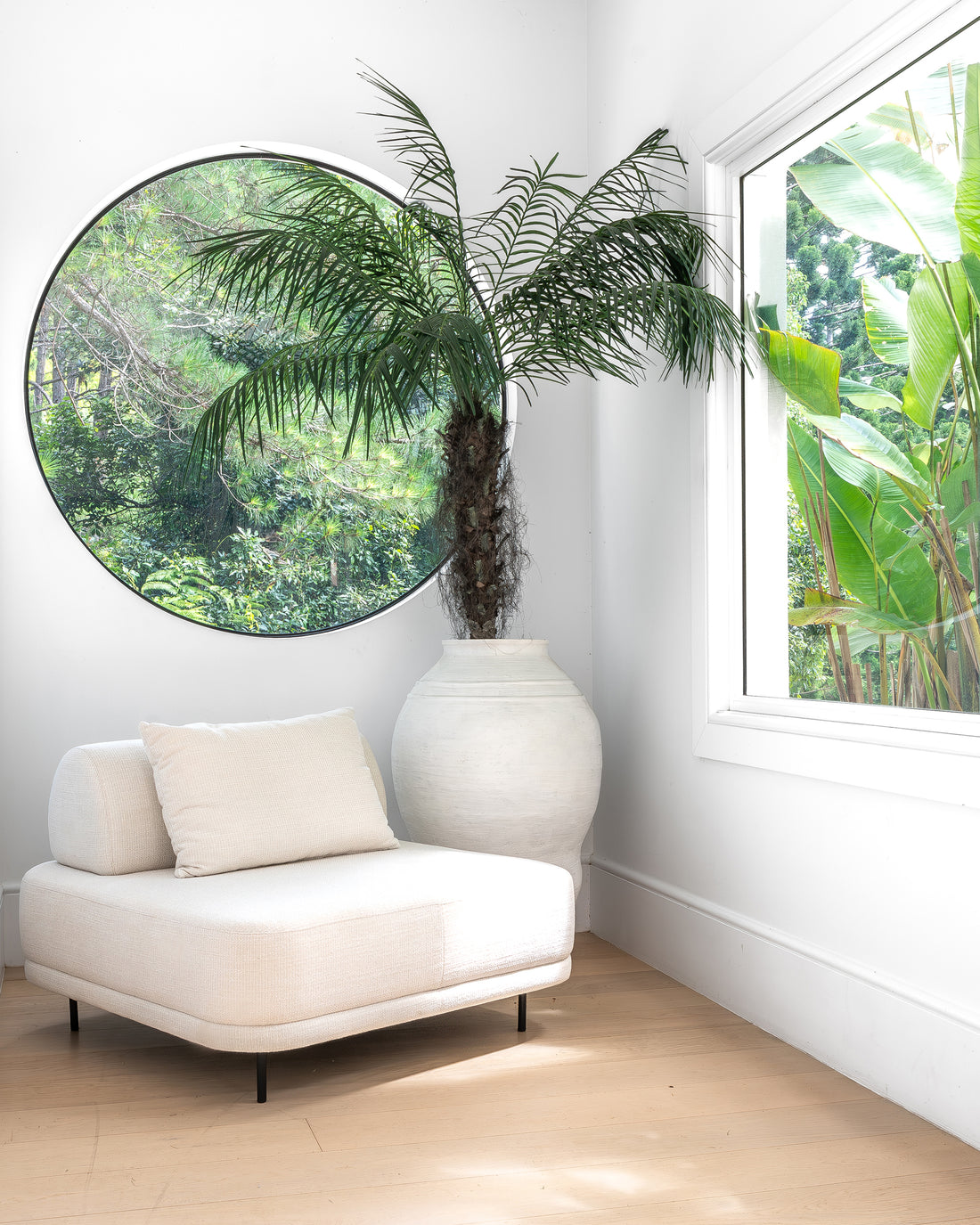 Modern white lounge chair styled with a textured cushion, positioned beside a grand white earthenware vase with lush greenery, reflecting in a large round window for a serene and organic corner