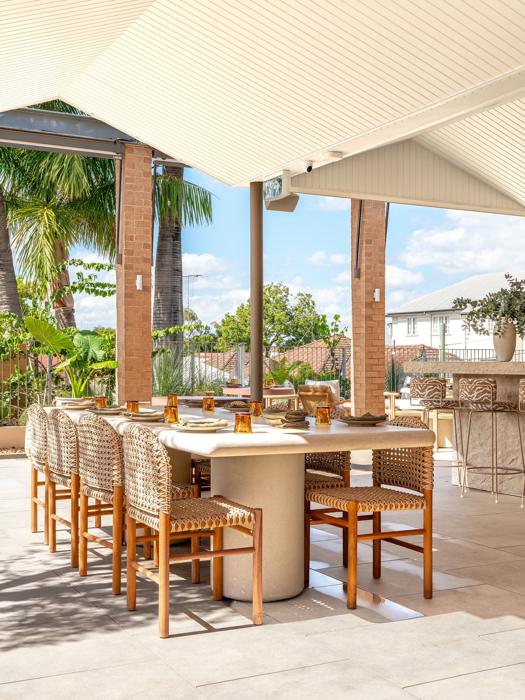 Mukalla dining table styled in an open rooftop bar setting with woven dining chairs warm brick columns and soft natural light creating a relaxed outdoor dining space