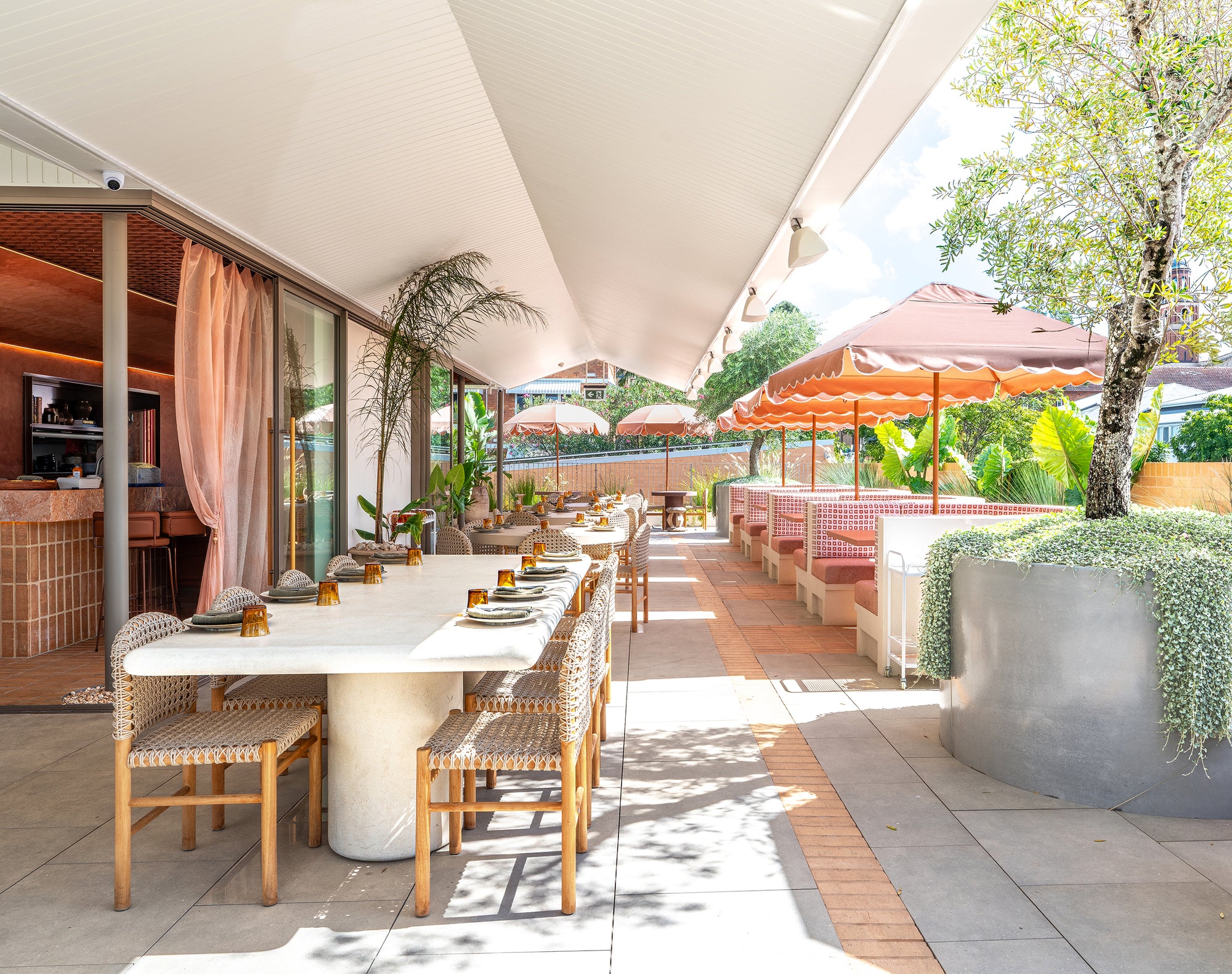 Mukalla dining table set along a sunlit rooftop terrace with woven chairs soft pink accents and lush greenery creating an inviting open air dining setting