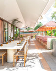 Mukalla dining table set along a sunlit rooftop terrace with woven chairs soft pink accents and lush greenery creating an inviting open air dining setting