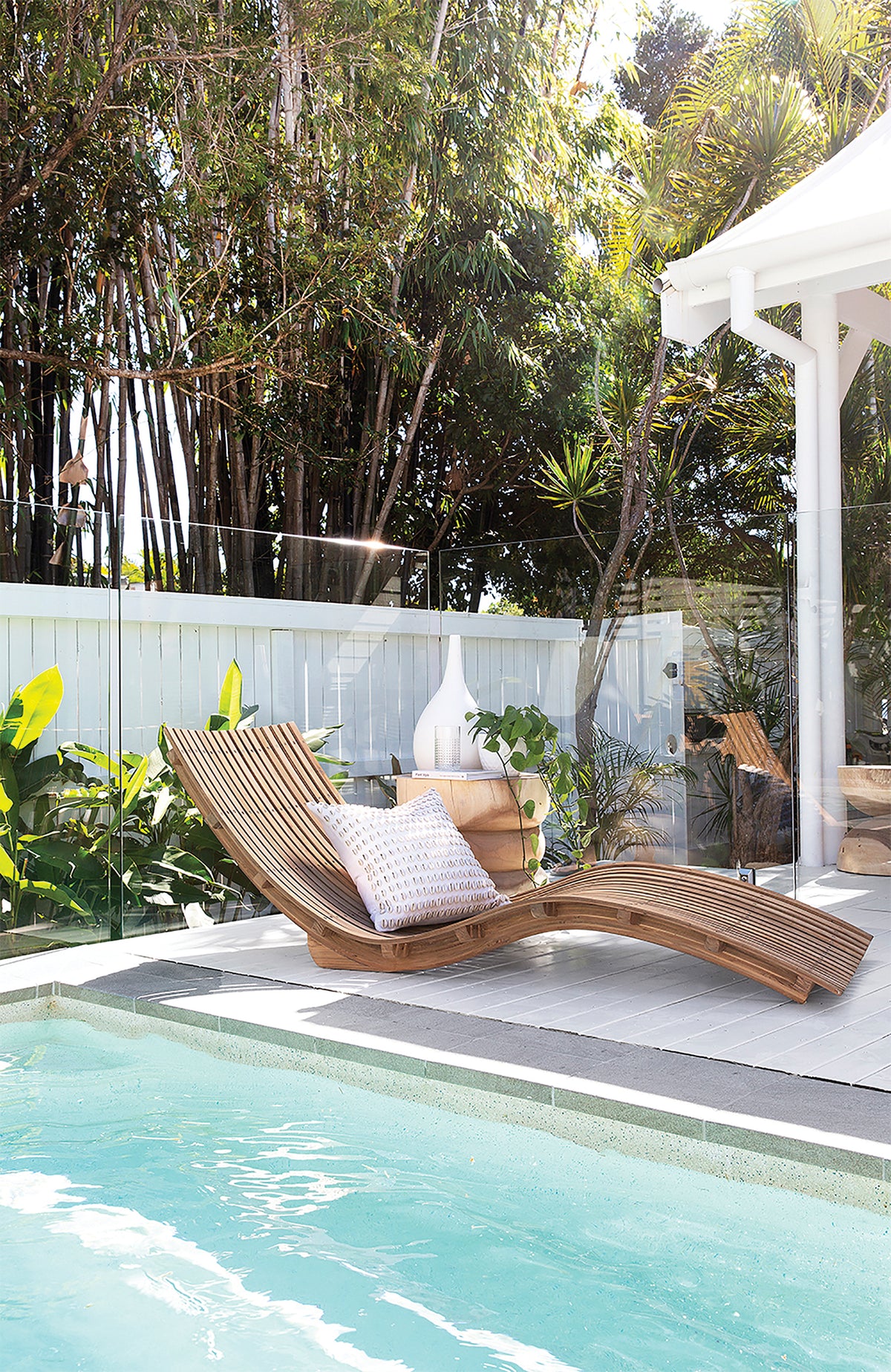 Mykonos Lounger with a curved slatted timber frame and white cushion, positioned beside a swimming pool surrounded by tropical greenery.