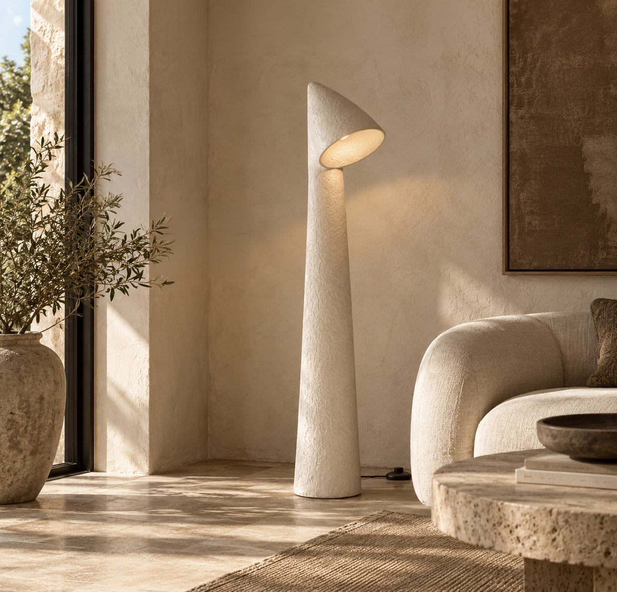 A modern sculptural floor lamp with a textured off-white finish stands in a sunlit minimalist living room next to a cream sofa and a large stone planter.
