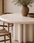 Close up of the Oman dining table featuring a rounded tabletop and sculptural fluted pedestal base styled with timber dining chairs in a warm neutral interior.