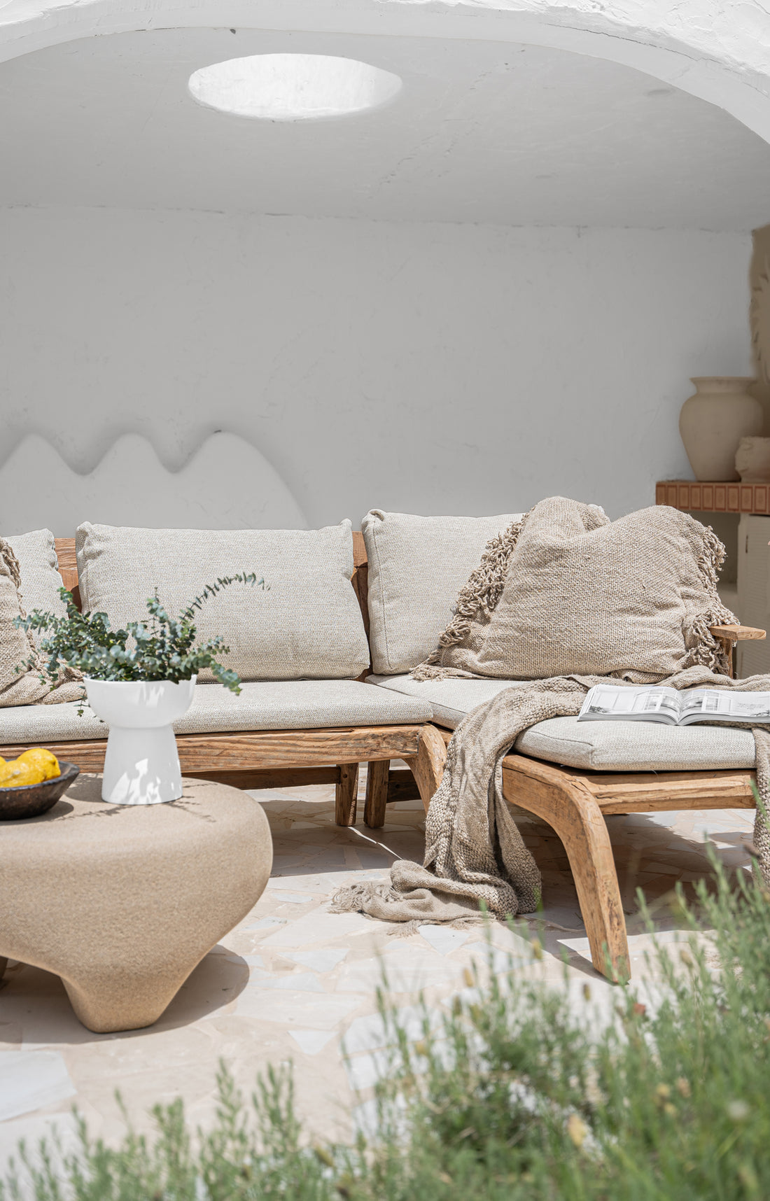 A beautifully curated outdoor lounge setting featuring a handcrafted teak sofa with plush neutral cushions, textured throw blankets, and organic décor, creating a relaxed resort-style ambiance.