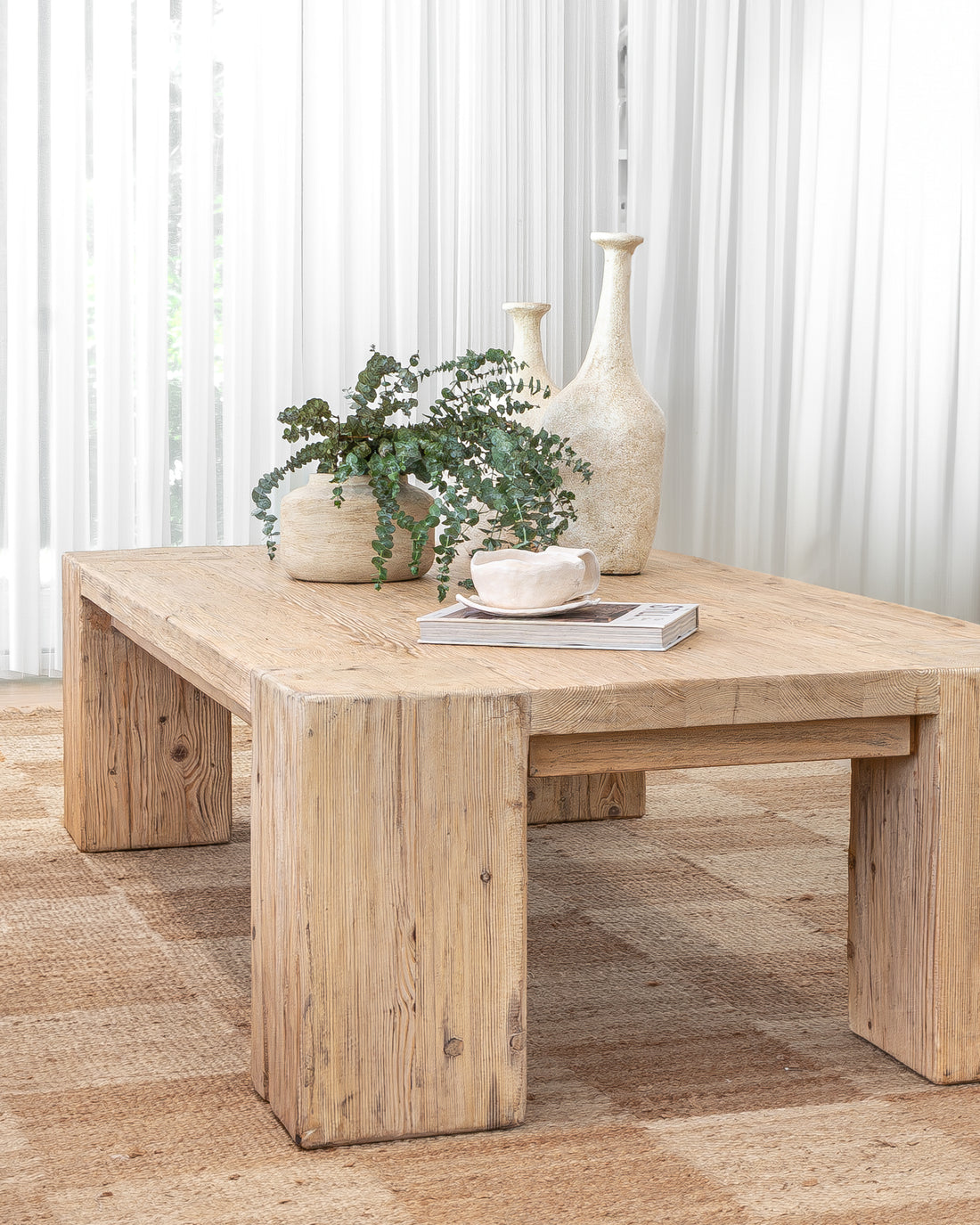 Uniqwa Collections a rustic reclaimed wood coffee table styled with textured ceramic vases, lush greenery, a sculptural cup, and a design magazine, set against soft white curtains for a tranquil and organic living space