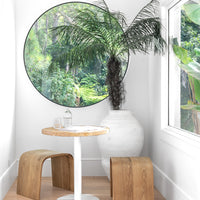 Round bistro table with a timber top and white pedestal base, paired with curved wood stools in a serene breakfast nook, complemented by a large round window and a white earthenware vase with lush greenery