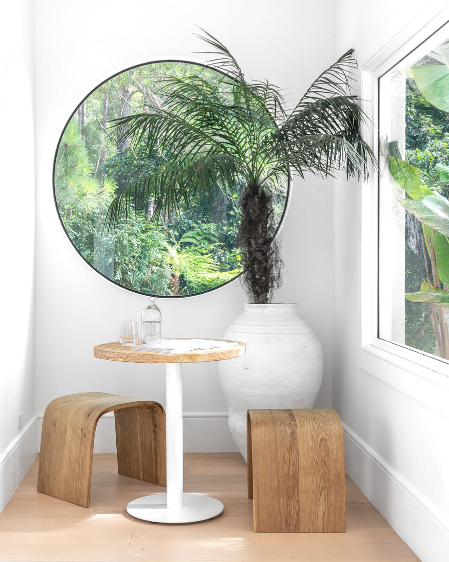 Round bistro table with a timber top and white pedestal base, paired with curved wood stools in a serene breakfast nook, complemented by a large round window and a white earthenware vase with lush greenery